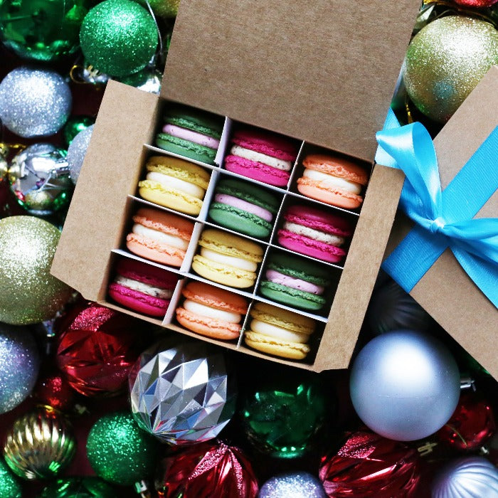 A box of 12 festive Christmas macarons on a pile of baubles.
Img Description: An open brown box of 12 colourful festive macrons, with a second brown box with a blue ribbon to the left of it. Both boxes are sat on a pile of Christmas baubles.