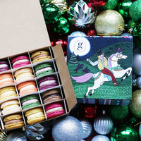 A range of 12 Christmas macarons in a festive box on top of a pile of Christmas baubles. 
Img Description: A range of 12 festive-flavoured macarons in a Christmas-themed box, on the box, is an illustration of a man wearing a top hat riding a horse through a field with Christmas trees in. The box is sat atop a pile of Christmas baubles. 