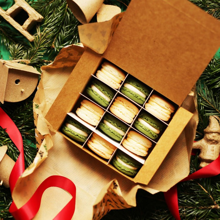 A box of 12 green and white festive macarons, laying atop some wrappig paper and a pile of pine branches.
Img Description: An open brown box of 12 green and white festive macrons, sat on top of some brown paper and a pile of pine branches.